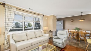 Un salon comprenant un canapé, un fauteuil, une table basse en verre, une table à manger avec des chaises, et une vue sur la rue grâce à de grandes fenêtres.