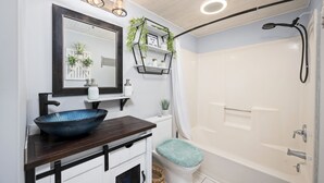 A bathroom with a large mirror, a wooden vanity, a blue bowl sink, a white toilet, a shower with a handheld showerhead, and shelves with plants and candles.