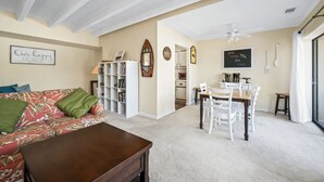 Una sala de estar con un sofá, una mesa de centro y una estantería. Hay una zona de comedor con una mesa y sillas. La habitación tiene un ventilador de techo y una ventana.