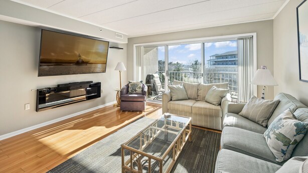 A modern living room with a flat-screen TV, two sofas, a glass coffee table, and a large window offering a view of the sea and buildings.