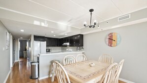 A modern kitchen with dark cabinets, a white countertop, and a dining area with a wooden table and white chairs.