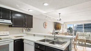 A modern kitchen with dark cabinets, a white countertop, a dishwasher, and a sink. There is a dining area with a table and chairs visible through an open door.