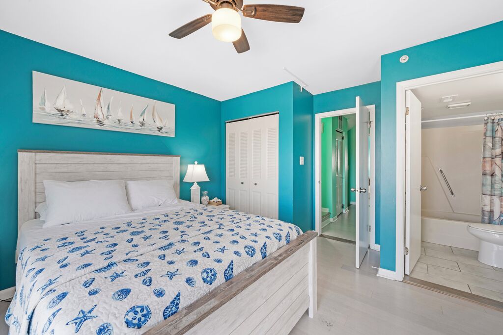 Ein Schlafzimmer mit Bett, Deckenventilator, einer Tür zu einem Badezimmer und einer Wand mit einem Seebild.