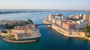 A coastal cityscape with historic fortifications, modern buildings, and a bridge connecting two areas. - Centralissimo e luminosissimo appartamento intero con Open Space. (Taranto)