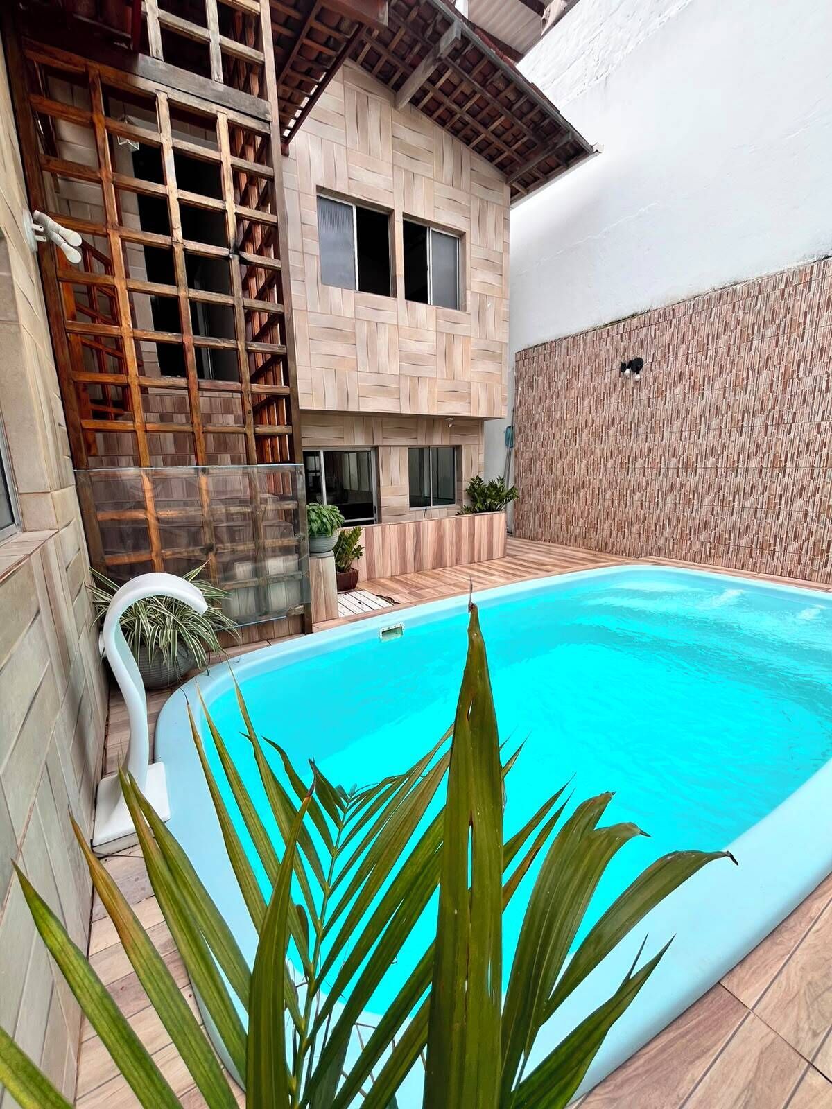 A swimming pool with a wooden deck, a modern house with a patterned facade, and a small garden area.