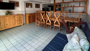 Ampla sala de estar com sofá, cadeiras de madeira, televisão e área de cozinha com armários e geladeira.
