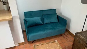 Un sofá color turquesa con dos cojines, una mesa de madera y un tapete con estampado de espiga sobre un suelo de baldosas.