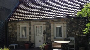 Un edificio de piedra con techo de tejas, dos ventanas, una puerta y un pequeño patio con una mesa y sillas.