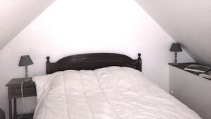 Una cama individual en una habitación con techo inclinado, dos mesitas de noche con lámparas y un estante con libros en la pared.