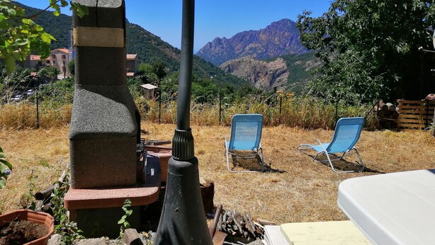 Une terrasse avec un barbecue en pierre, deux chaises bleues et une vue sur les montagnes.