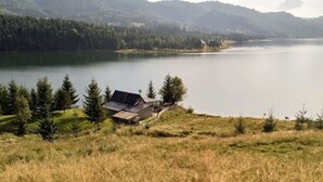 En stuga vid sjön med ett sluttande tak, omgiven av ett skogsområde och med berg i bakgrunden.