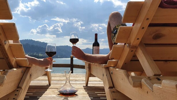 Dos personas sentadas en sillas de madera mirando un lago con montañas al fondo.