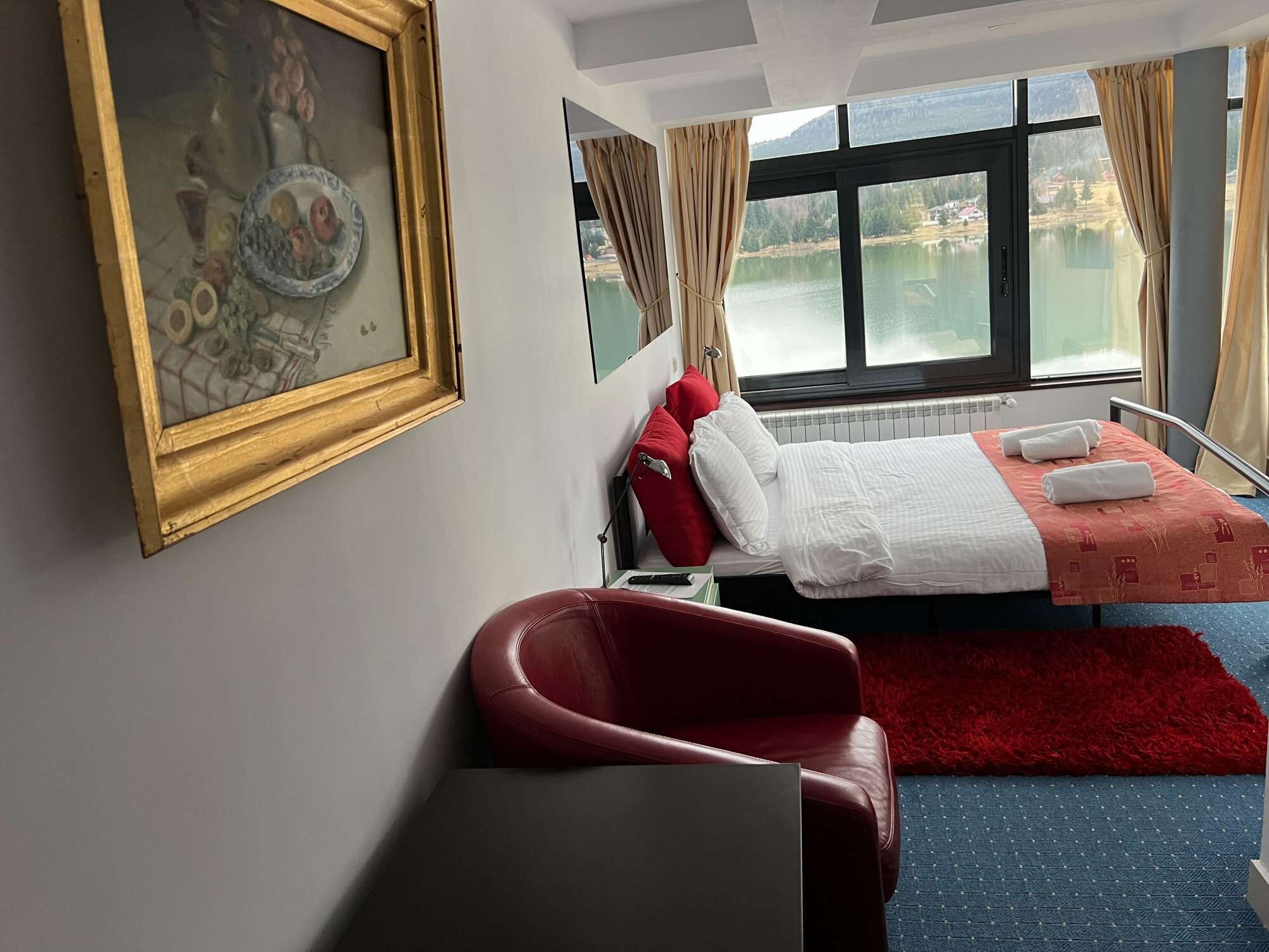 Habitación de hotel con cama, una silla roja, un cuadro y ventana con vistas a un lago.
