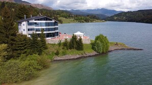 Un moderno edificio de dos pisos con amplios ventanales que dan a un lago, rodeado de vegetación y montañas.