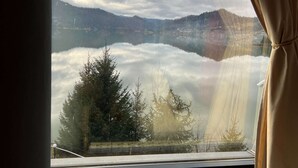 Vista desde una ventana con cortina, mostrando un lago, árboles y montañas a lo lejos.