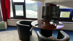Una habitación con un ventanal grande, un televisor sobre una mesa de madera y dos sillas negras.