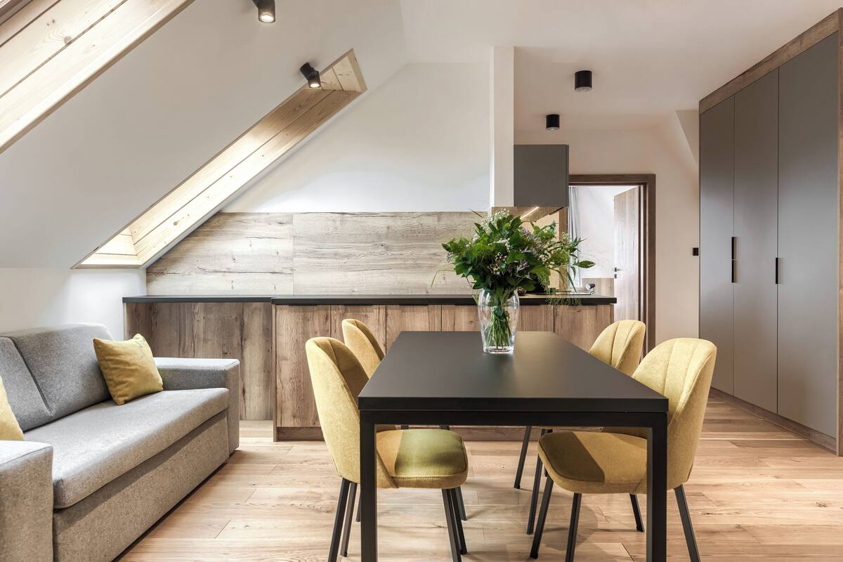 Eine moderne Küche mit einer Wand in Holzoptik, ein Essbereich mit einem schwarzen Tisch und gelben Stühlen sowie ein graues Sofa mit einem gelben Kissen.