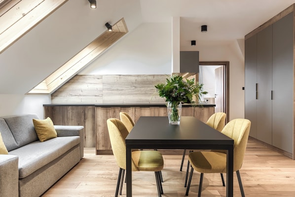 Una cocina moderna con una pared de acento de madera, un área de comedor con una mesa negra y sillas amarillas, y un sofá gris con un cojín amarillo.