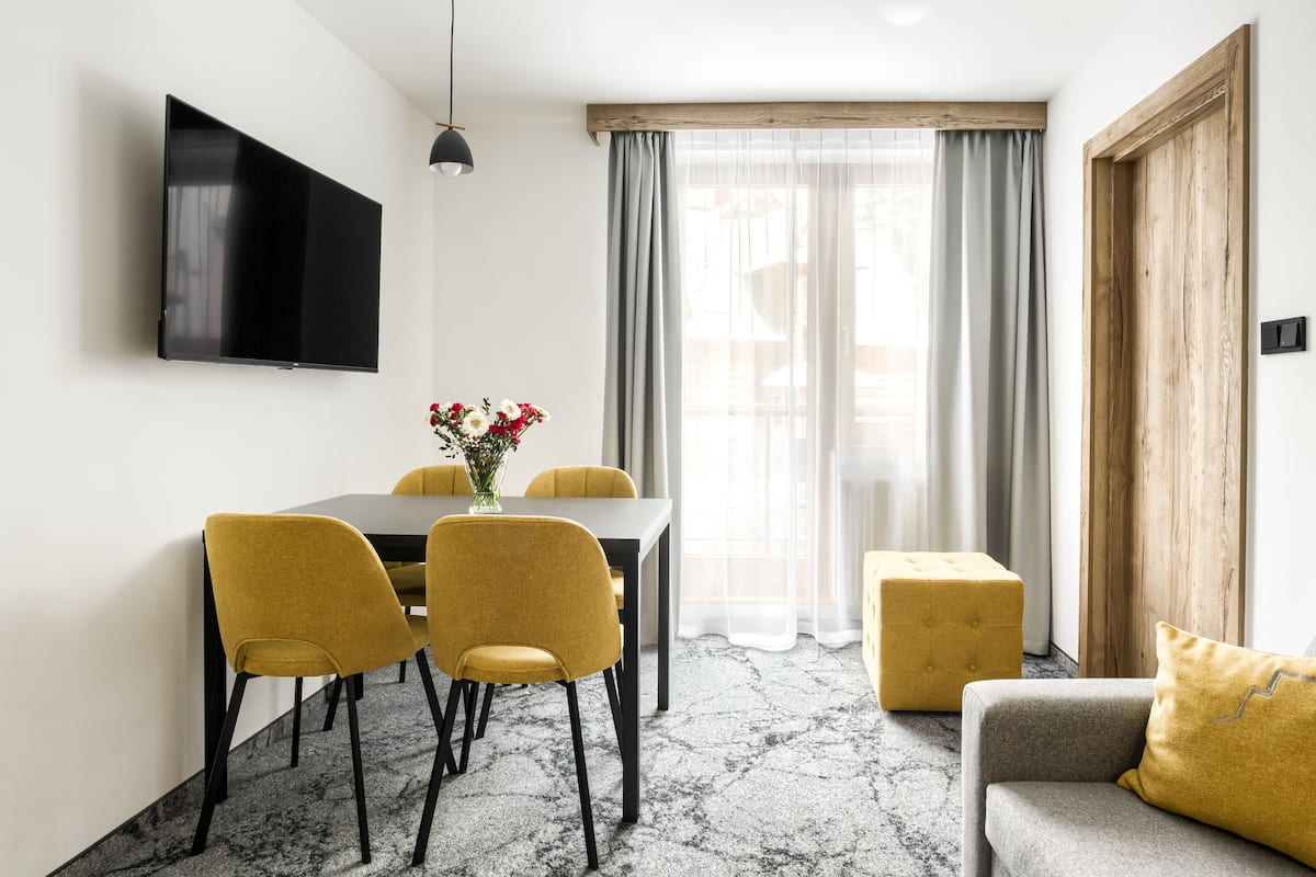 Ein modernes Zimmer mit Esstisch, vier Stühlen, einer Couch, einem an der Wand montierten Fernseher und einem fenster mit Vorhängen.