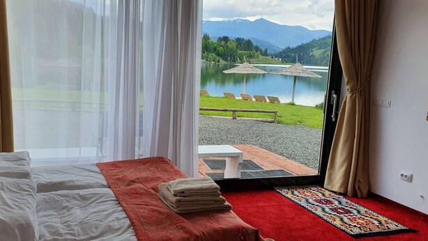 Une chambre à coucher avec un lit, un tapis rouge et une vue sur un lac et des montagnes, visible par une porte ouverte.