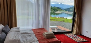 A bedroom with a bed, a red rug, and a view of a lake and mountains through an open door.
