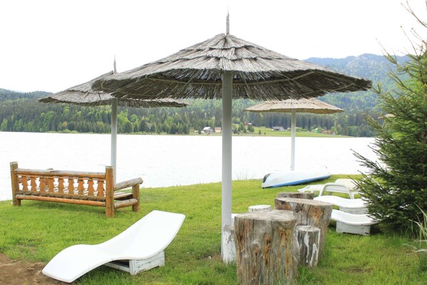 A lakeside setting with a wooden bench, a lounge chair, a small boat, and a thatched umbrella.