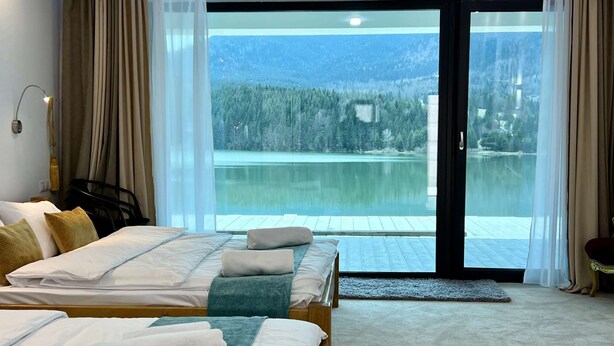 Habitación de hotel con dos camas, vista a un lago y un bosque, y puerta corrediza de vidrio.