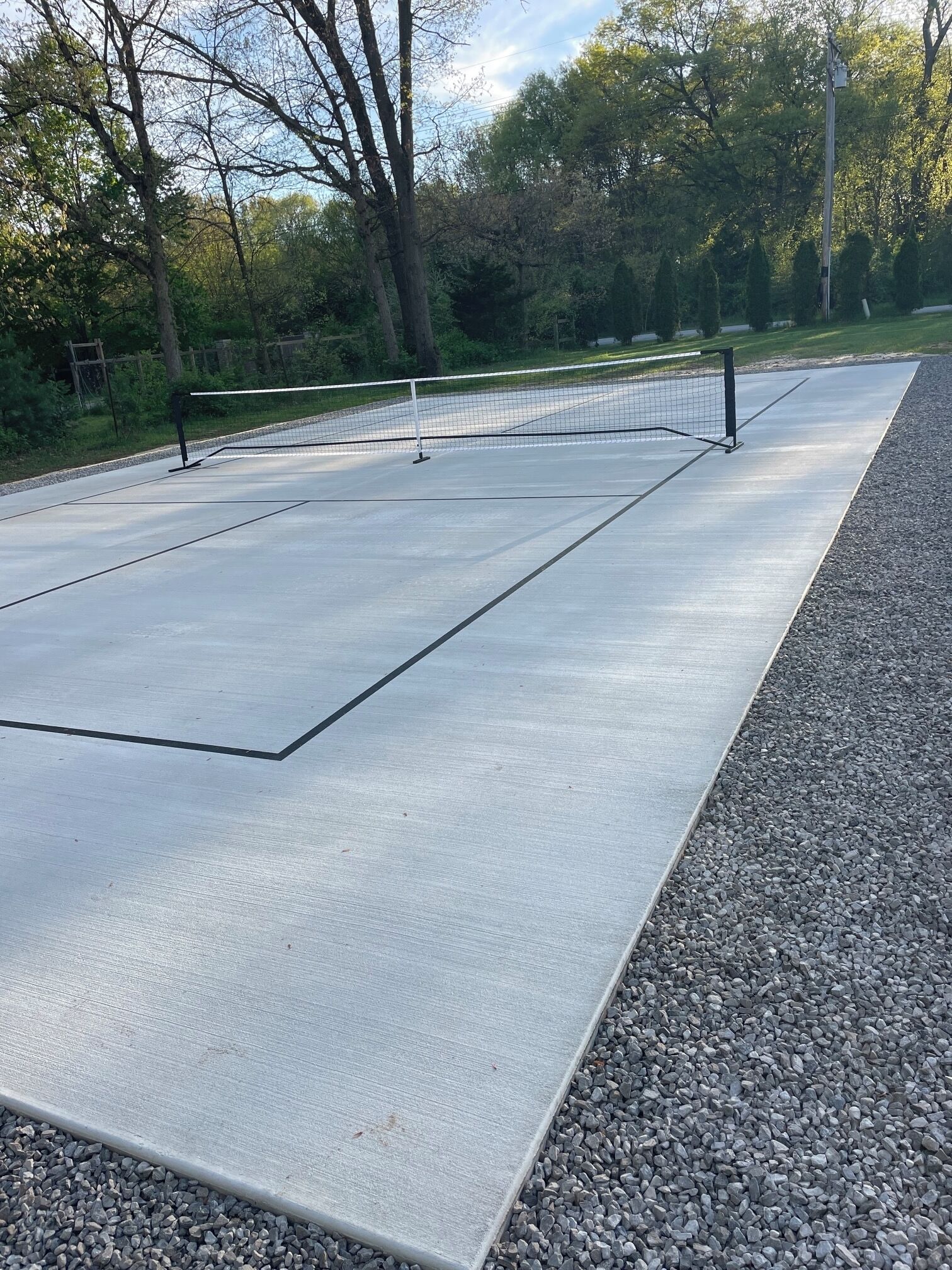 A tennis court with a net, surrounded by trees and a gravel path.
