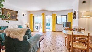 A living room with a sofa, a dining table with chairs, a TV, and yellow curtains. - Casa Felix with pool (Albufeira)