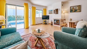 A living room with a TV, sofa, and a fireplace. There is a balcony with yellow curtains and a pool in the background. - Casa Felix with pool (Albufeira)