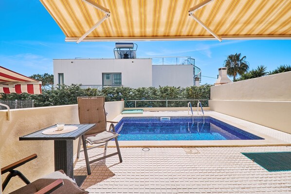 A rooftop pool area with a chair and table, a pool, and a building in the background.