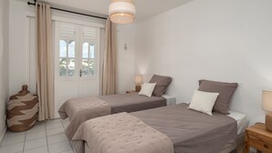 Ein Schlafzimmer mit zwei Betten, einem Fenster mit Vorhängen, einer Nachttischlampe und einem gewebten Korb.