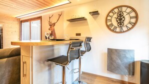 A modern kitchen with a wooden ceiling, a bar area with two stools, a large clock, and a deer head decoration. - Le Cerf des Alpages - Appt cosy pour 2 (La Giettaz)