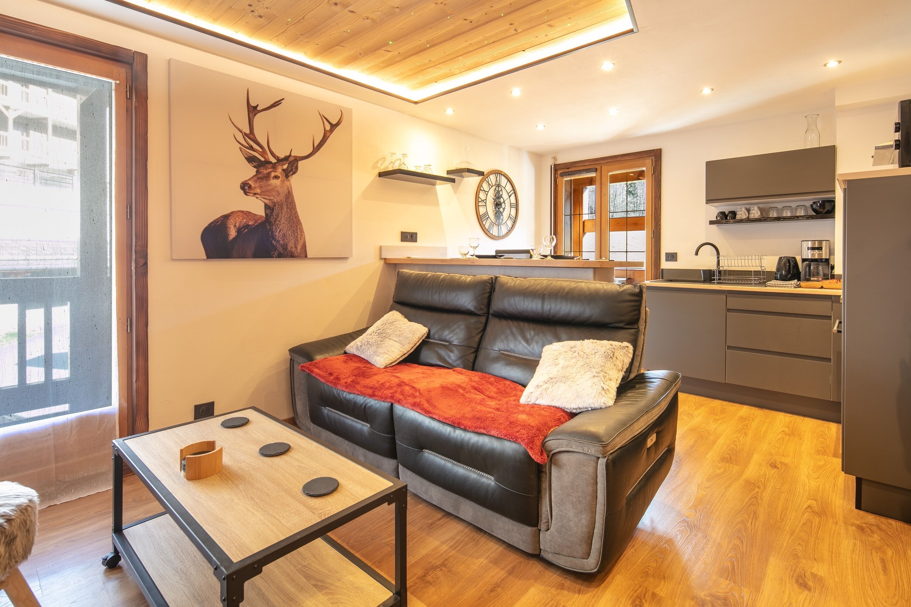 Una sala de estar moderna con un sofá de cuero, una mesita de centro y una mesa de comedor. Hay una zona de cocina con un fregadero y armarios. Un cuadro de un ciervo adorna la pared, y se puede ver un reloj.