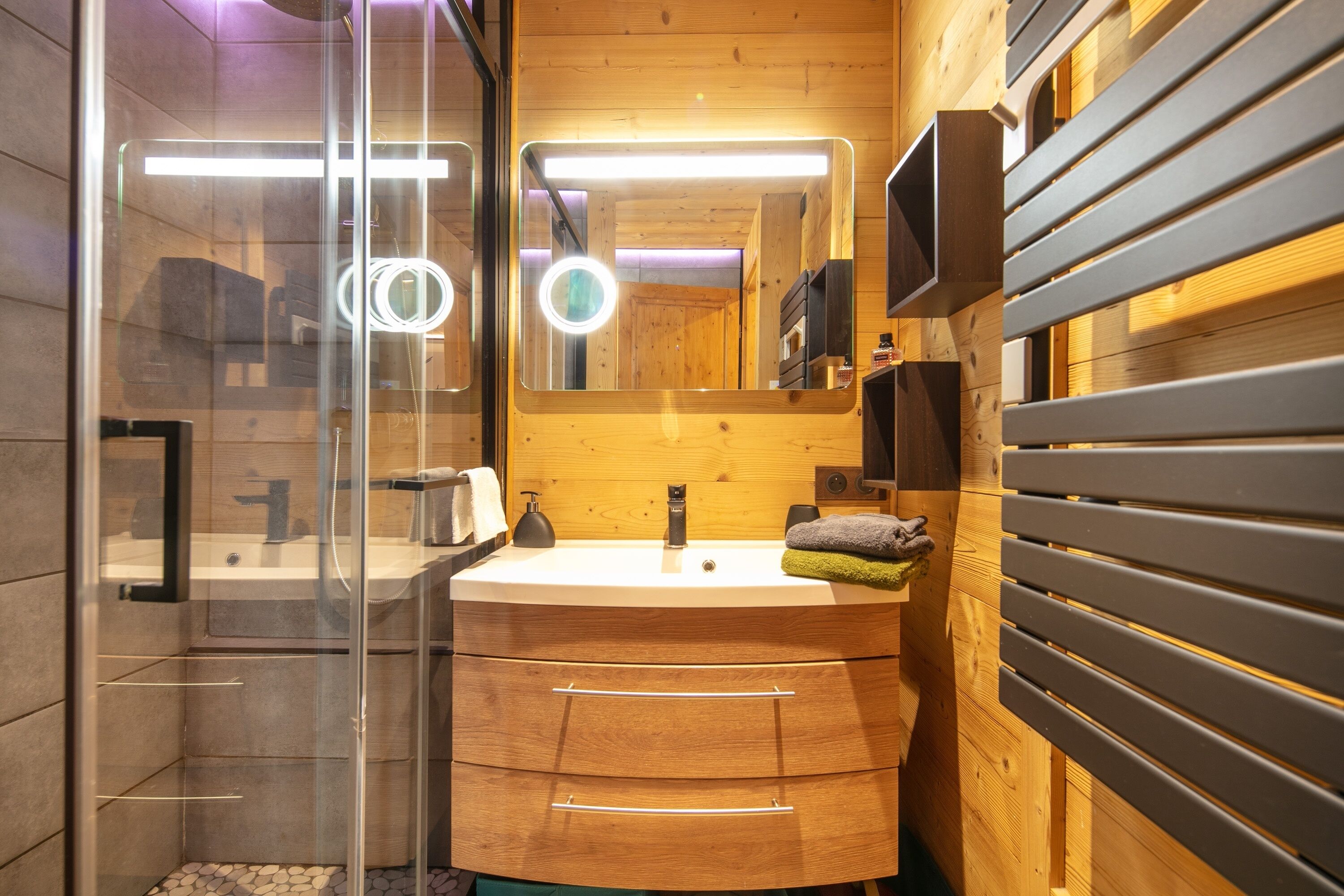 Une salle de bain moderne avec un meuble-lavabo en bois, une cabine de douche en verre et un miroir doté d’un anneau lumineux.