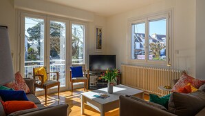 Ein geräumiges Wohnzimmer mit großem Fernseher, bequemen Sitzgelegenheiten und großen Fenstern mit Blick auf die Umgebung.
