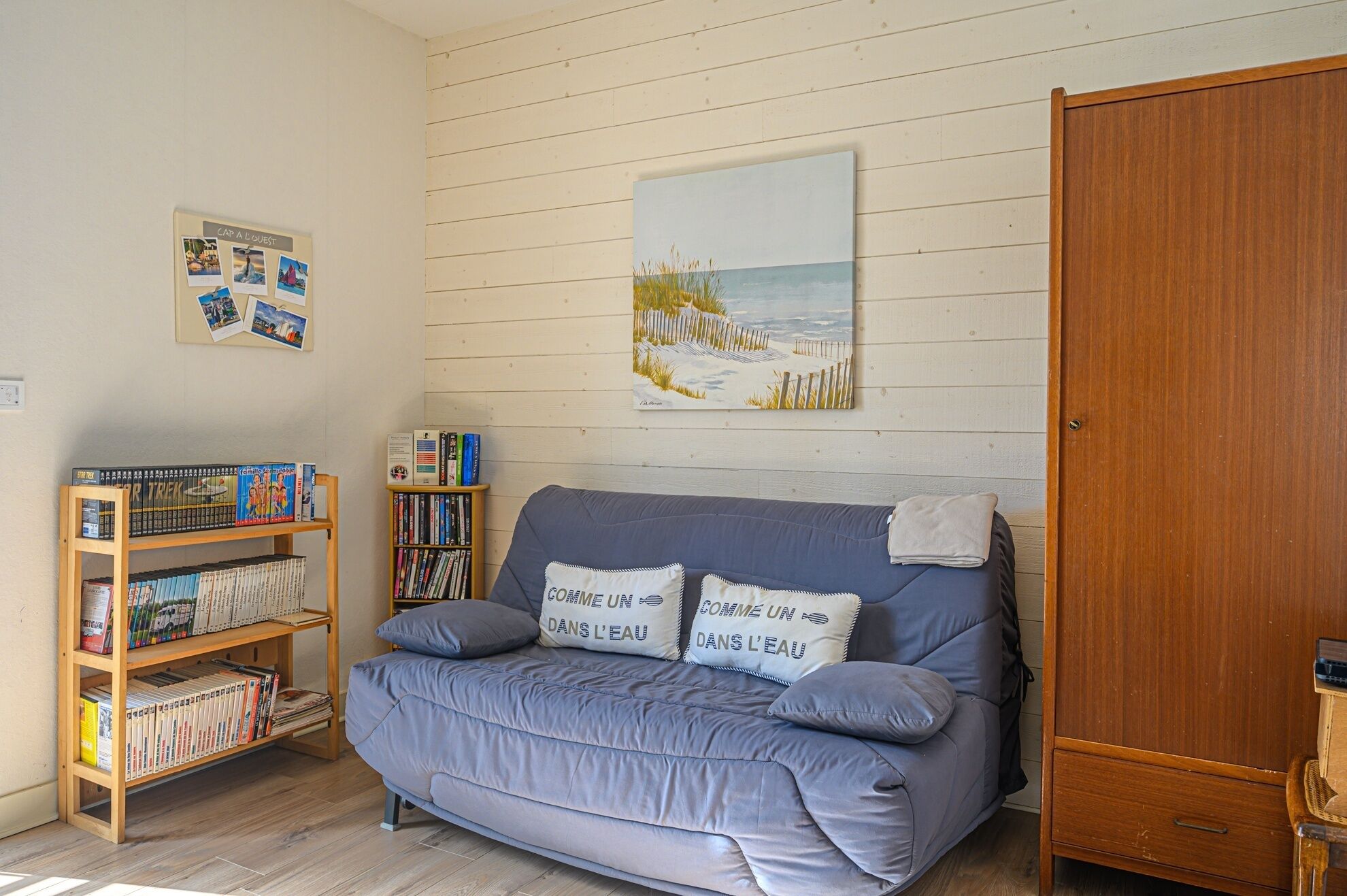 A room with a grey sofa, wooden bookshelves, a wooden wardrobe, and a beach-themed painting on the wall.