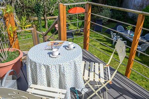 Une table ronde recouverte d’une nappe à motifs floraux, une chaise en métal et une plante en pot, le tout placé sur une terrasse en bois offrant une vue sur un jardin et un espace de détente extérieur.