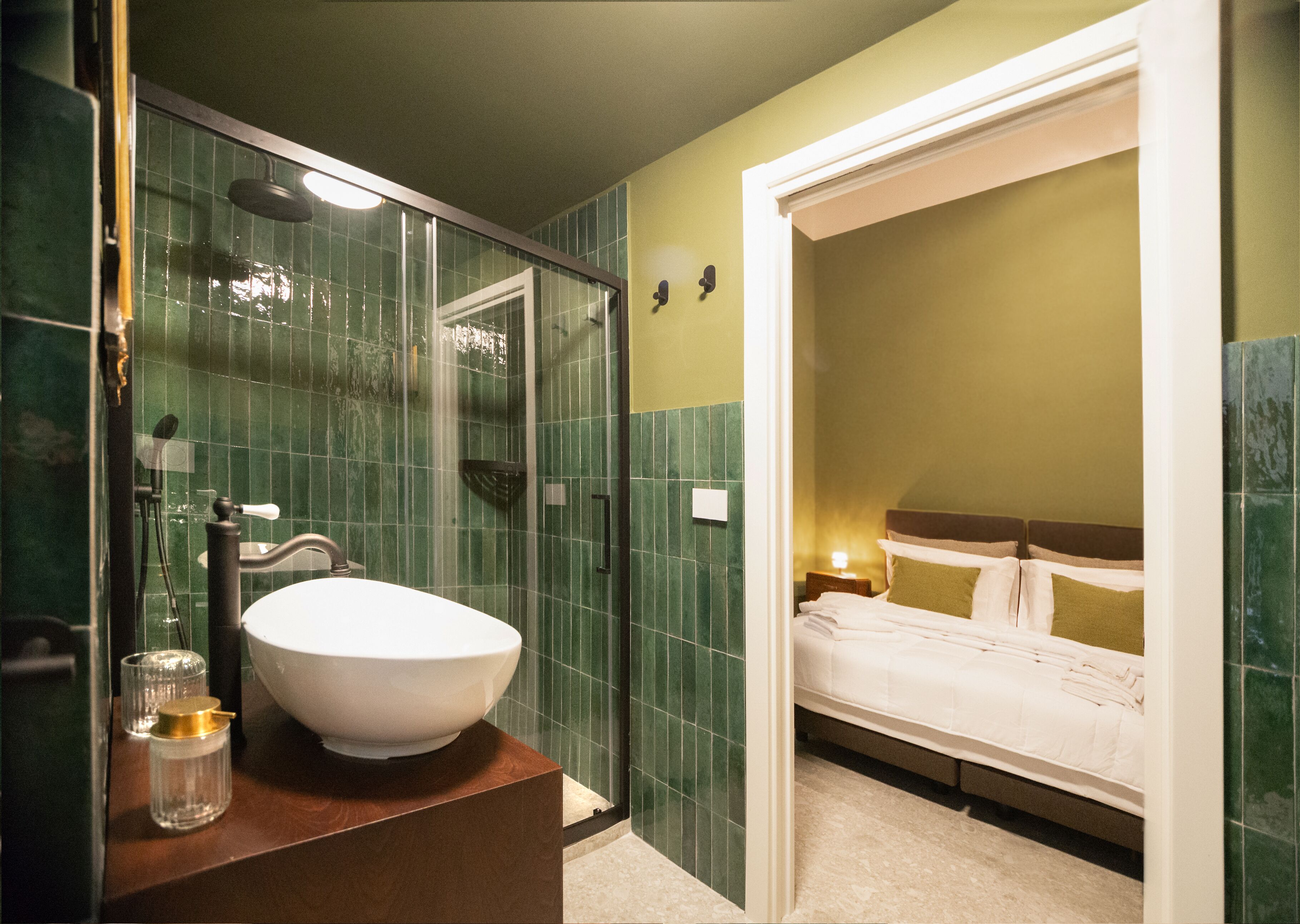 A bathroom with a large white sink, a glass shower enclosure, and a bed visible through an open doorway.