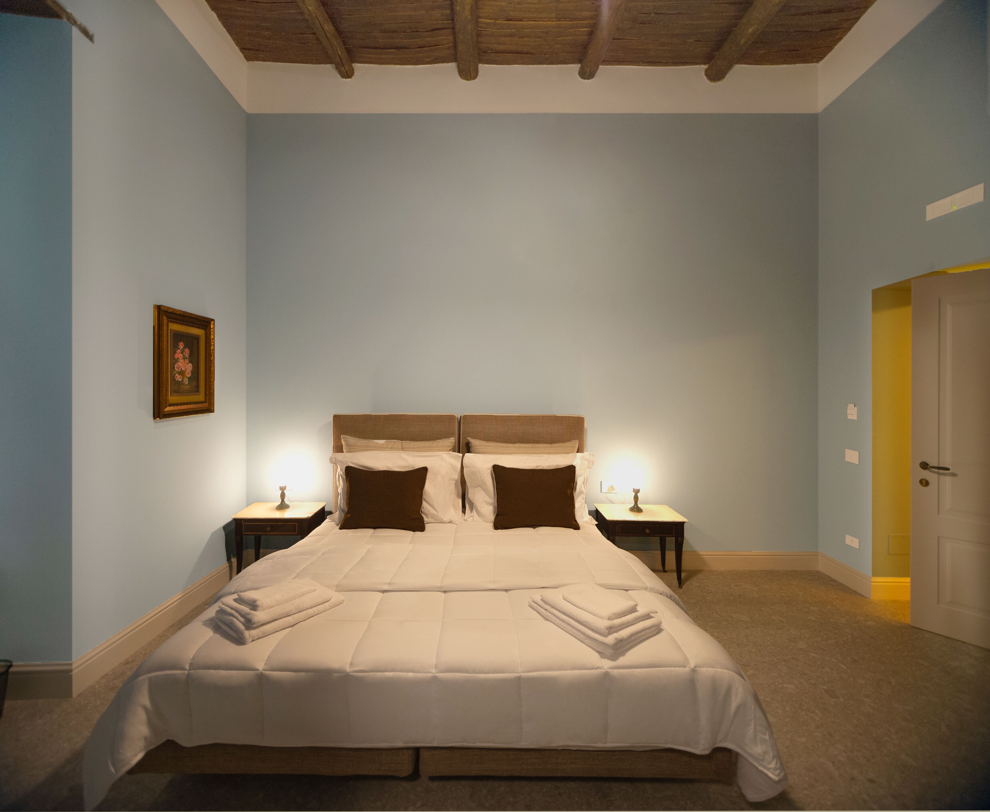 A bedroom with a large bed, two bedside tables, a framed picture on the wall, and a door to the right.