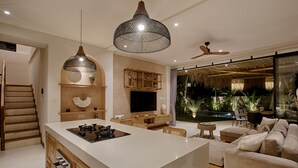 A modern kitchen with a central island, wooden cabinetry, and a large TV. There is a dining area with a sofa and a small table. A staircase is visible on the left, and a large window provides a view of a pool and greenery. - Villa Nueva Expectacular. Vive Bingin Beach!! Trona, Cuna, Bañera Gratis (Bali)