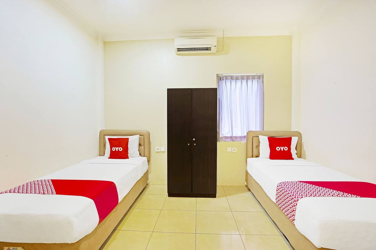 A hotel room with two single beds, a wardrobe, an air conditioning unit, and a window with curtains.