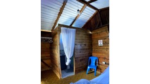 A rustic wooden cabin with a single room featuring a bed, a blue chair, and a shower area. - Villa Cruz Hotel & Restaurant Windsurf Fan Eco Rm (buen hombre)