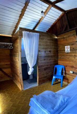 A rustic wooden cabin with a single room featuring a bed, a blue chair, and a shower area.