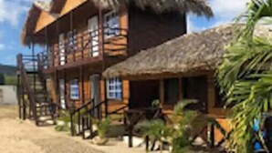 A wooden building with a thatched roof, a staircase leading to a balcony, and a sandy ground with small plants. - Villa Cruz Hotel & Restaurant Windsurf Fan Eco Rm (buen hombre)