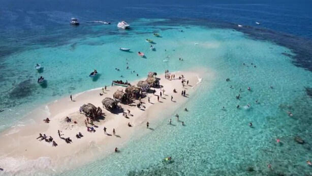 Vista aérea de una playa con aguas turquesas cristalinas, gente nadando y tomando el sol, y embarcaciones a lo lejos.