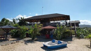 Un edificio frente a la playa con un amplio patio abierto, rodeado de palmeras y un bote en la arena.
