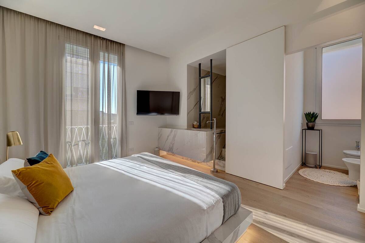 A modern hotel room with a large bed, a TV, a bathroom with a sink and mirror, and a balcony with a view.