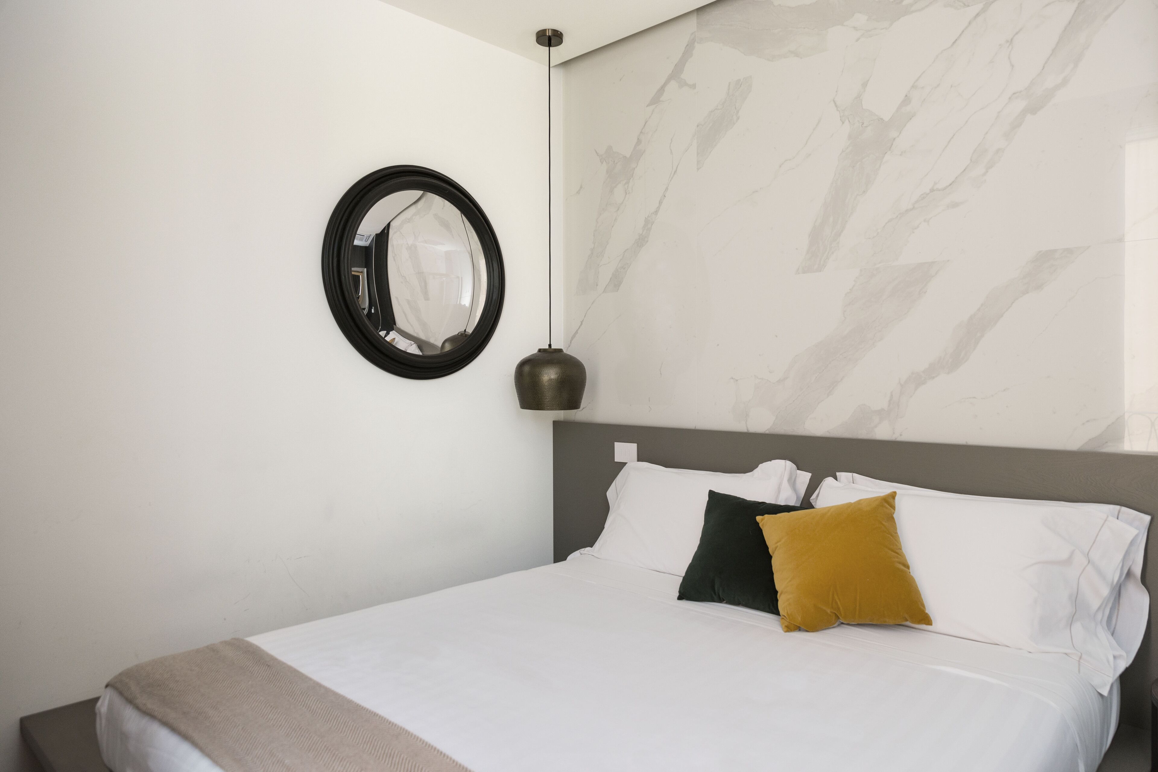 A neatly made bed with white linens and two pillows, a round mirror on the wall, and a pendant light.