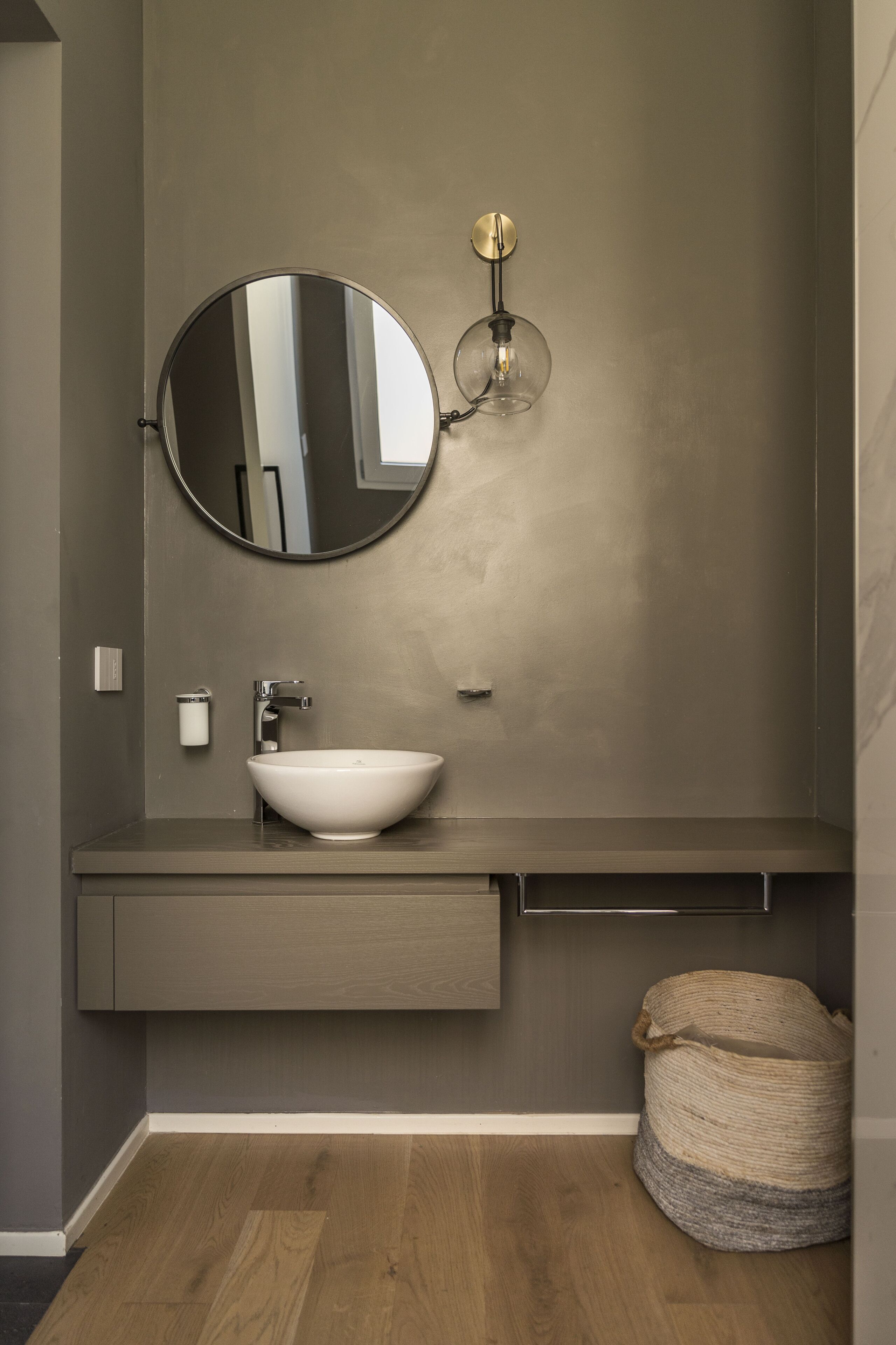 A modern bathroom with a round mirror, a white sink, a towel rack, and a small storage cabinet.